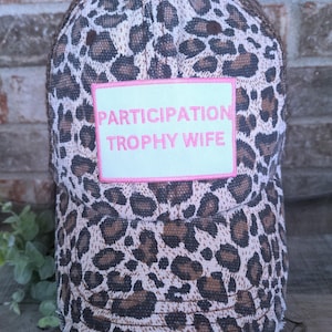 May include: A baseball cap with a leopard print pattern in shades of brown and tan. The cap features a mesh back and a patch with the words "PARTICIPATION TROPHY WIFE" in pink lettering.