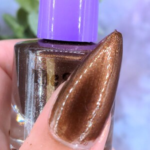 May include: A bottle of brown nail polish with a purple cap. The nail polish is being tested on a long, almond-shaped nail. The nail polish has a metallic, shimmering finish.