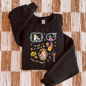 Könnte beinhalten: Schwarzes Sweatshirt mit farbenfrohen Illustrationen einer Prinzessin, eines Frosches, eines Pferdes und anderer Märchenmotive. Das Design umfasst ein Schloss, eine Laterne und florale Elemente. Das Sweatshirt ist aus weichem Material.