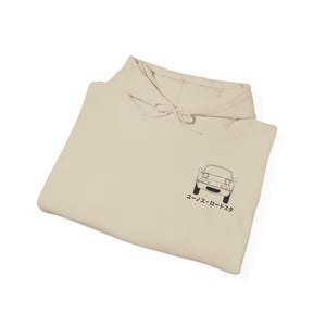 May include: A beige hoodie with a drawstring hood. The front of the hoodie features a black line drawing of a car and Japanese text. The hoodie is folded and appears to be made of a soft material.
