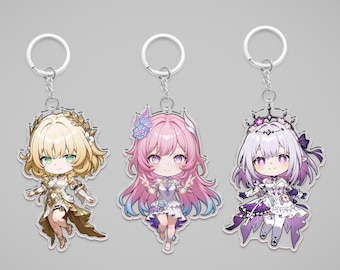 May include: Three anime-style keychains featuring chibi characters. The keychains have silver rings and chains. The characters have blonde, pink, and lavender hair, and wear detailed outfits in gold, white, and purple. The background is a solid gray.