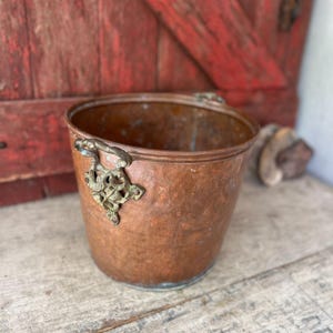 May include: A round, hammered copper bucket featuring a wide, open top and a flat base. The exterior surface displays a warm, metallic copper tone. Two ornate, decorative brass handles are attached to the sides, showcasing intricate metalwork.