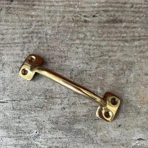 May include: A polished brass handle with a curved bar and rectangular mounting plates with two holes each. The handle is set against a weathered, gray wooden surface.