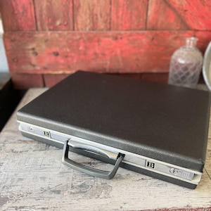 May include: A vintage black briefcase with silver trim and a gray handle. The briefcase has a textured surface and features the letters "R" and "B" on the sides. The briefcase is closed and appears to be in good condition.
