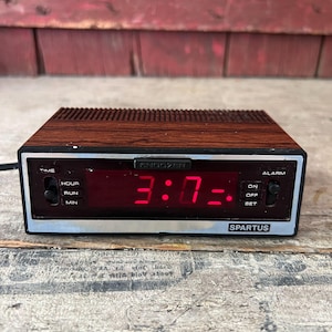 May include: A rectangular digital alarm clock with a faux wood grain top and sides. The front panel features a black display with bright red digital numbers showing 3:07, a silver trim, and the brand name Spartus. It includes a Snoozer button.