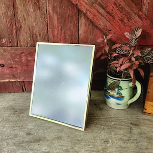 May include: A rectangular picture frame with a thin, metallic gold-toned border and a glass front. The frame is positioned upright, displaying a neutral, light gray interior surface. The design is simple, featuring clean lines and a polished finish.
