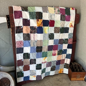 May include: A patchwork quilt constructed from numerous square fabric pieces in a variety of colors and patterns, including solid white, blue, green, red, and brown. The squares are joined with light blue yarn ties at each corner intersection.
