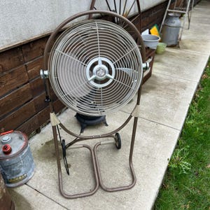 May include: A vintage brown and beige oscillating fan with a metal cage and blades. The fan has a circular design and is mounted on a brown metal frame with wheels for easy movement. The fan is designed for cooling and air circulation.