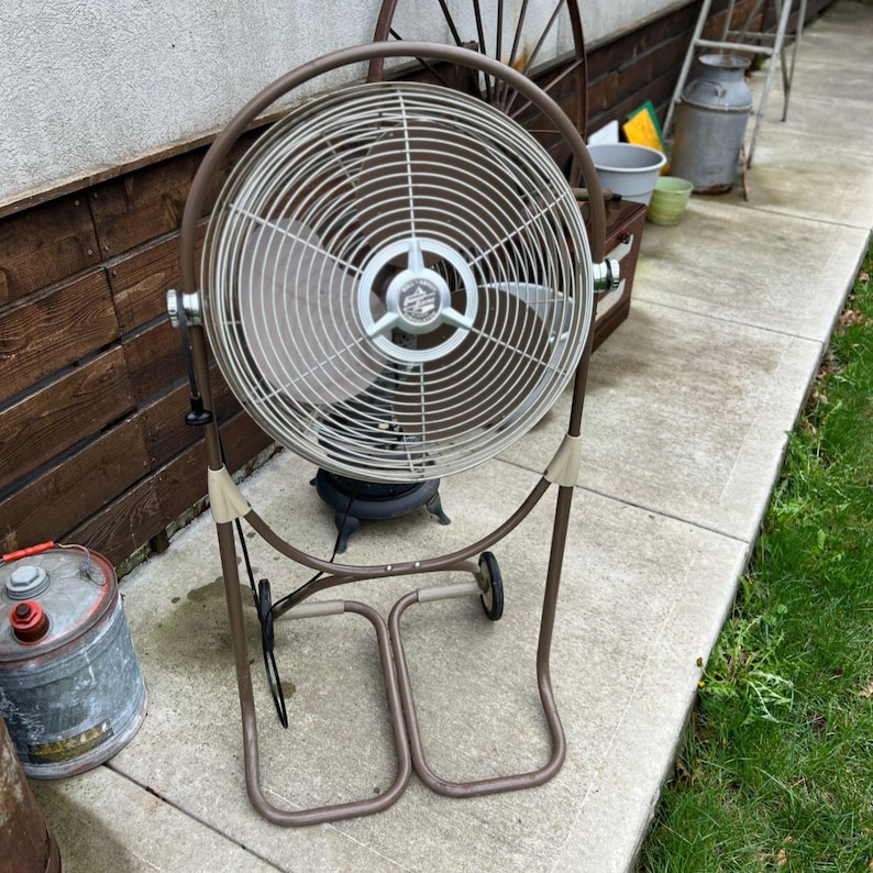 May include: A vintage brown and beige oscillating fan with a metal cage and blades. The fan has a circular design and is mounted on a brown metal frame with wheels for easy movement. The fan is designed for cooling and air circulation.