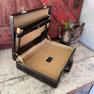 May include: A dark brown hard-shell briefcase with a tan interior lining. The open case features a document divider, pen loops, and a small black rectangular label with white text. It includes gold-toned metal hinges, latches, and a handle.