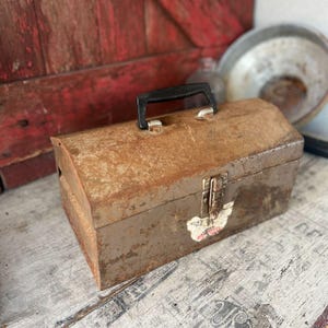 May include: A vintage, weathered metal toolbox with a black handle and a latch. The toolbox has a rusty, brown finish with a small sticker on the front. The toolbox is sitting on a distressed wooden surface.