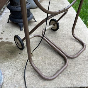 May include: A brown metal plant stand with two small wheels for easy movement. The stand has a curved design and a black cord is attached. The stand is on a concrete surface.
