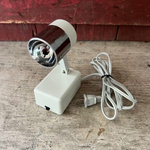 May include: A white spotlight lamp with a chrome-finished cylindrical head and a rectangular base. The lamp features an adjustable hinge, a black power switch on the side of the base, and a white electrical cord with a two-prong plug attached.