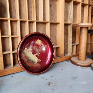 May include: A round, red lacquered tray with a gold rim. The tray features a landscape design with a mountain, pagoda, and trees in gold and cream colors. The tray is likely vintage and decorative.