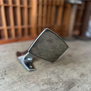 May include: A silver-toned metal cuff bracelet with a large, square, textured center piece. The bracelet has a curved design and is set against a wooden surface.