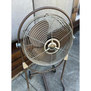 Pu&ograve; includere: Ventilatore oscillante vintage marrone e argento con gabbia circolare e supporto con ruote. Il ventilatore ha una gabbia in metallo e un emblema centrale. Il supporto &egrave; marrone con dettagli beige.