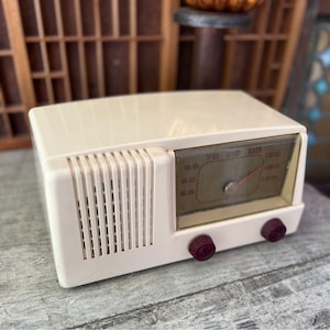 May include: Cream-colored vintage radio with a rectangular shape. The radio features a clear dial with red markings and a red needle. Two maroon knobs are on the front. The radio has a speaker grill on the left side.