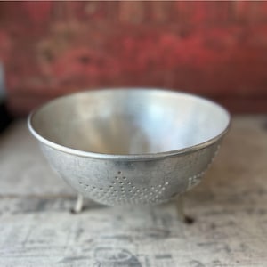 May include: A vintage, silver-colored metal colander with small feet and numerous drainage holes. The bowl-shaped kitchen tool is designed for straining and rinsing food items. The colander is set against a rustic, blurred background.