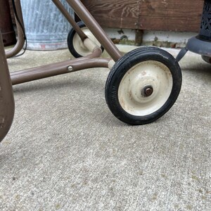 May include: Close-up of a vintage brown metal cart with two wheels. The wheels have black tires and off-white centers. The cart's frame is a muted brown color, and the wheels are attached to the frame with metal hardware. The cart appears to be outdoors.