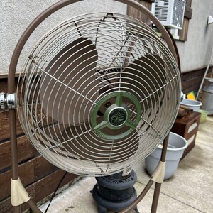 May include: A vintage brown and silver oscillating fan with a circular cage and blades. The fan has a green and silver center with the text "Roll About Emerson Electric Circulator". The fan is on a brown stand.