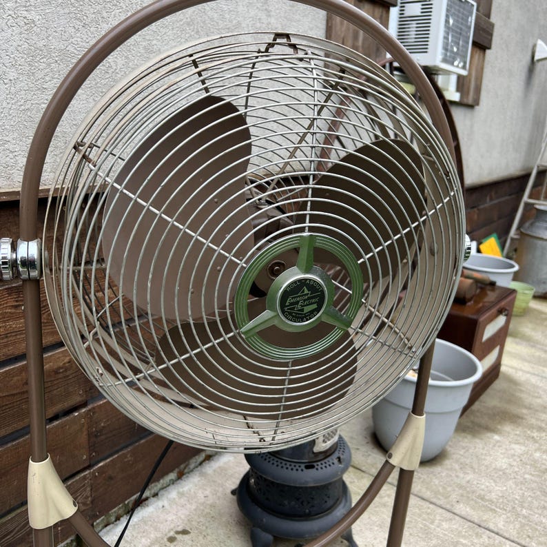 May include: A vintage brown and silver oscillating fan with a circular cage and blades. The fan has a green and silver center with the text "Roll About Emerson Electric Circulator". The fan is on a brown stand.