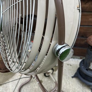 May include: An antique, beige and brown oscillating fan with a chrome knob. The fan has a cage-like design with curved blades and a brown stand. The fan is a vintage appliance.