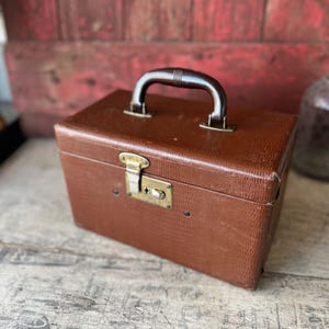 May include: A vintage brown faux-leather travel case with a textured surface. The case has a dark brown handle and a brass-colored clasp. The case is rectangular and appears to be in good condition.
