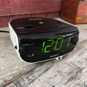 May include: Black and white Sony Dream Machine CD clock radio with a digital green display showing 12:01 PM. The clock has buttons for alarm, track, and sleep functions. The CD player is on top.
