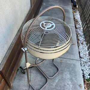 Pu&ograve; includere: Un ventilatore oscillante vintage bianco sporco e marrone, con una gabbia circolare e una maniglia curva. Il ventilatore ha una base marrone con ruote. La gabbia metallica ha un design circolare. Per la circolazione dell'aria.