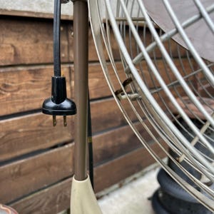 May include: Close-up of a vintage oscillating fan with a black power cord and plug. The fan has a metal cage and a brown and cream-colored base. The background is a wooden fence.