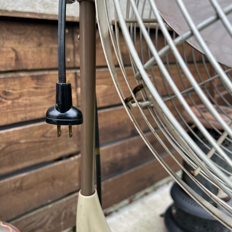 May include: Close-up of a vintage oscillating fan with a black power cord and plug. The fan has a metal cage and a brown and cream-colored base. The background is a wooden fence.