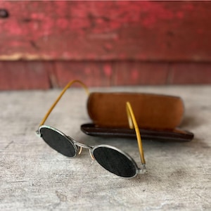 May include: Vintage oval sunglasses with dark lenses and thin, silver frames. The temples are a light brown color. A brown leather glasses case sits in the background.