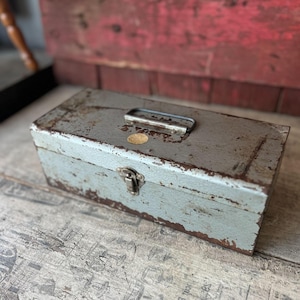 May include: A weathered, rectangular metal toolbox with a handle and clasp. The gray paint is chipped, revealing rust-colored metal underneath. A small, round sticker is affixed to the front. The toolbox is likely used for storing tools.