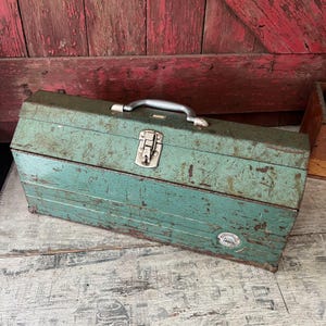 May include: Vintage teal metal toolbox with a silver handle and latch. The toolbox shows signs of wear, with scratches and a weathered appearance. A small, circular emblem is visible on the front. The toolbox is rectangular and has a hinged lid.