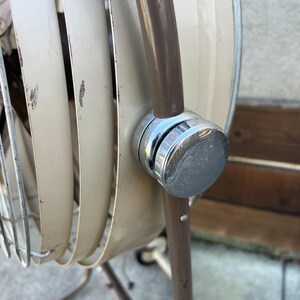 Pu&ograve; includere: Primo piano di un ventilatore oscillante vintage beige e marrone. Il ventilatore ha una manopola cromata e una gabbia di metallo. Le pale del ventilatore sono beige e il telaio &egrave; marrone. Il ventilatore &egrave; in stile retr&ograve;.