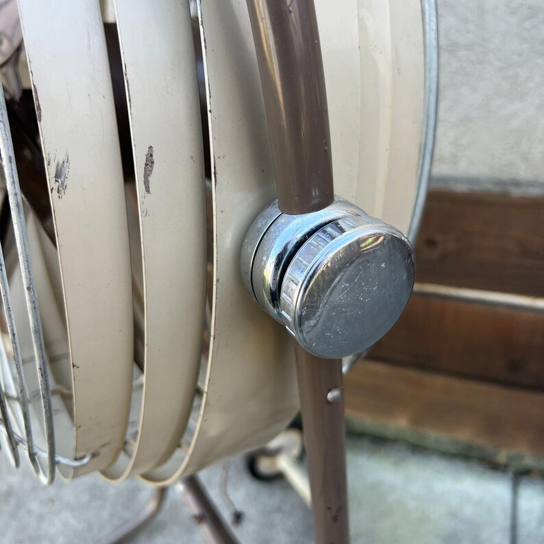 Pu&ograve; includere: Primo piano di un ventilatore oscillante vintage beige e marrone. Il ventilatore ha una manopola cromata e una gabbia di metallo. Le pale del ventilatore sono beige e il telaio &egrave; marrone. Il ventilatore &egrave; in stile retr&ograve;.