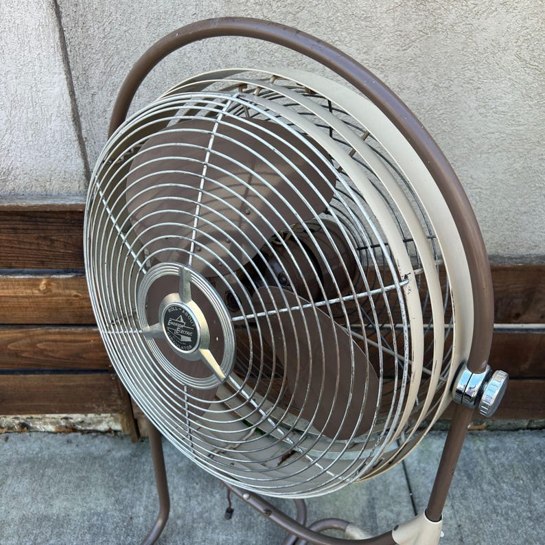 Pu&ograve; includere: Un ventilatore oscillante vintage marrone e crema. Il ventilatore ha una gabbia circolare con un telaio marrone e una protezione per le pale color crema. La base e il supporto sono anch'essi marroni. Ha una manopola di controllo cromata e una maniglia.