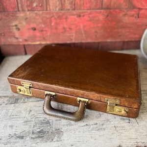 May include: A vintage brown leather briefcase with brass hardware. The briefcase has a handle and two latches. The leather shows signs of wear, suggesting it is an antique or has been well-used. The briefcase is closed.