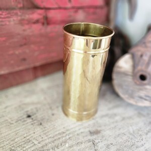 May include: A tall, cylindrical brass vessel with a polished, reflective surface. The container features a smooth, unadorned exterior with a slightly flared rim at the top and a reinforced base, showcasing a warm, metallic gold-toned finish throughout.