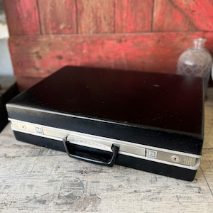 May include: A black vintage briefcase with silver trim and a black handle. The briefcase has a rectangular shape and a lock on the side. The brand name "PPM" is visible on the front. The briefcase is sitting on a wooden surface.