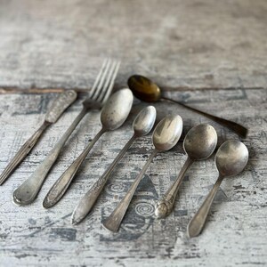 May include: An assortment of vintage silverware, including spoons, a fork, and a knife, arranged on a weathered, light-colored wooden surface. The spoons vary in size and design, with some showing ornate detailing. The silverware has a tarnished, antique appearance.