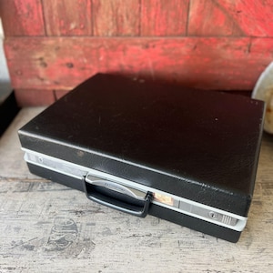 May include: A black Samsonite briefcase with silver trim and a black handle. The briefcase has a rectangular shape and a textured surface. The case is closed and appears to be in good condition.