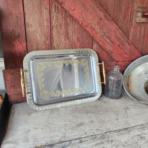 May include: A rectangular silver-toned metal serving tray with gold-colored handles and decorative trim. The tray has a gold scroll design in the center. A glass bottle with a wire cage and a metal bowl are also visible.
