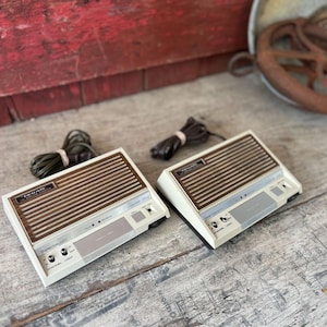 May include: Two vintage Realistic Selecticom intercom units. Each unit has a cream-colored base with a brown, slatted top. The units have a series of buttons and dials, and a cord. The units are on a weathered wooden surface.