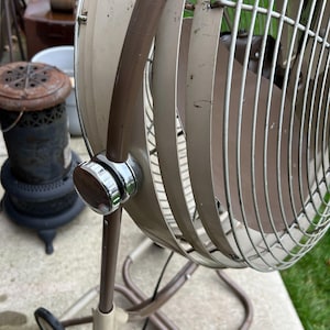 May include: Vintage beige and brown oscillating fan with a chrome and wood-grain control knob. The fan has a metal cage and blades, and is mounted on a stand with wheels. The fan is an antique, and shows signs of wear.