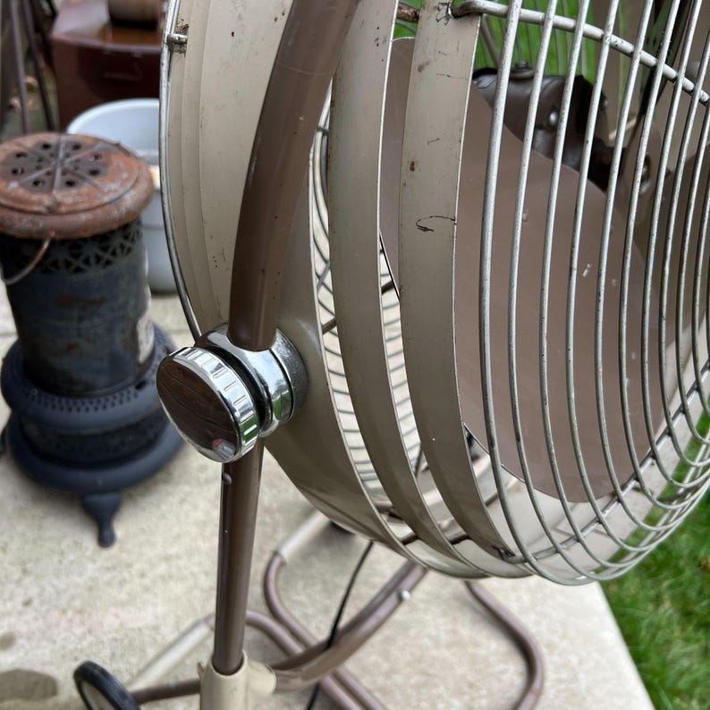 May include: Vintage beige and brown oscillating fan with a chrome and wood-grain control knob. The fan has a metal cage and blades, and is mounted on a stand with wheels. The fan is an antique, and shows signs of wear.
