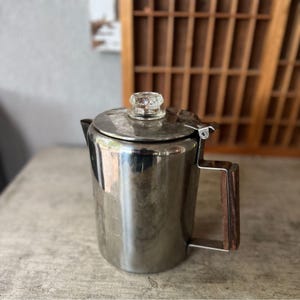 May include: A stainless steel coffee percolator with a clear glass knob on the lid and a dark wood handle. The vintage coffee pot is sitting on a light-colored surface.
