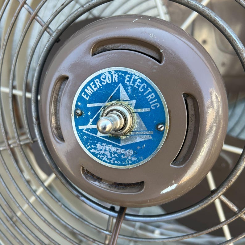 Pu&ograve; includere: Primo piano di un ventilatore Emerson Electric. Il ventilatore ha un alloggiamento motore marrone con un emblema blu e bianco. L'emblema recita "Emerson Electric" e presenta un design a stella e triangolo. Le pale del ventilatore sono racchiuse in una gabbia metallica.