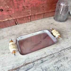 May include: A rectangular serving tray with a metallic finish and decorative handles. The handles feature a combination of cream-colored and amber-toned elements. The tray is set against a rustic wooden background.