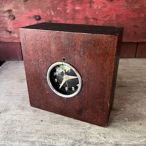 May include: A vintage wooden clock with a dark brown finish. The clock face is black with white numerals and hands, and is encased in a silver-colored metal frame. The clock is rectangular and appears to be made of solid wood.